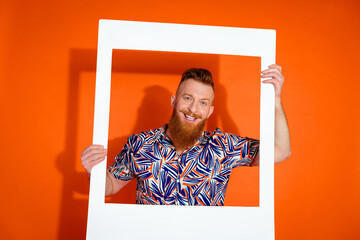 Portrait of toothy beaming man with red long beard wear print shirt posing in white frame isolated on vibrant orange color background