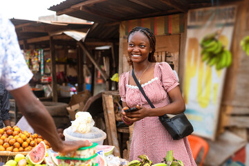 Confident Young Black African Saleswoman Assisting Client at Fruit Stand with Mobile Phone in Hand