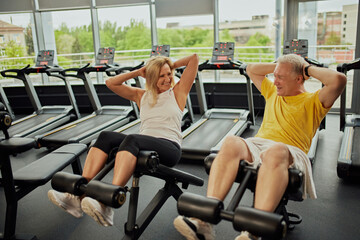 Senior couple, man and woman, laughs while doing abdominal exercises at modern gym for retirements....