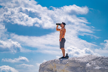 A man is standing on a rock and drinking water. The sky is cloudy and the man is wearing a hat