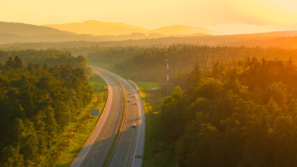 AERIAL, LENS FLARE: Morning light spills over hilly wooded landscape and highway. Trucks and cars travel along the scenic motorway leading through beautiful countryside bathing in golden sunlight.