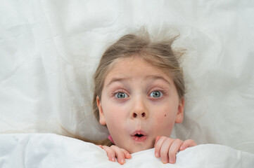 Top view of a grimacing little girl lying in bed. 