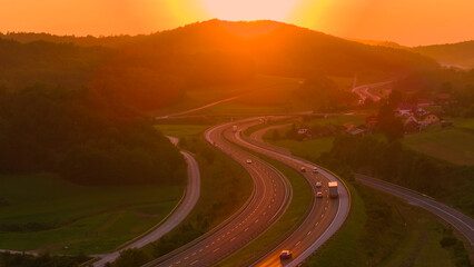 AERIAL, LENS FLARE: Morning rays peek over hill and reflect from driving vehicles. Sun rises over a highway winding through hilly countryside, casting long shadows and painting sky in orange shades.