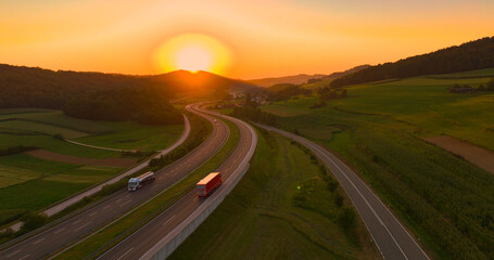 AERIAL, LENS FLARE: A stunning sunset over hilly landscape and winding highway. Calm evening and light traffic on the motorway passing green countryside colored in orange hue of setting summer sun.