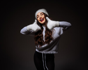 A woman in a white sweater and black pants is smiling and making a funny face. She is wearing a white hat and has red lips