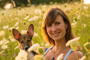 PORTRAIT, DOF: Cheerful lady poses with her young adopted puppy amidst lush blooming wildflowers. Woman holds her doggo as they sit surrounded by the beauty of a blossoming meadow in golden sunlight.