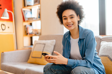Smiling African American woman holding mobile phone chatting, sitting on comfortable sofa