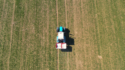 Agricultural tractor with trailer for organic fertilizers driving at rural field. Machine sprays saltpeter chemical from trailer onto land, fertilizing soil for best growth plant. Aerial view follow