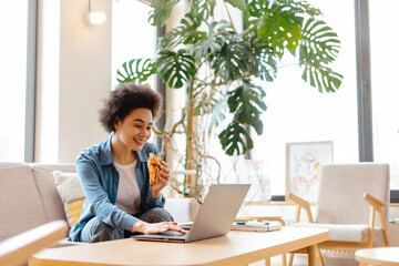 Happy attractive African American woman eating croissant, using laptop, working in cozy cafe