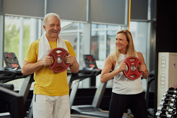 Fototapeta na wymiar Senior man and woman doing exercises, happily lifting weight plates in sunlit modern gym. Concept of sport, active seniors in modern life, healthy lifestyle, fitness centers, hobby, strength and power