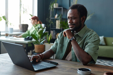 Portrait of smiling African American man using laptop at workplace in open office decorated with...