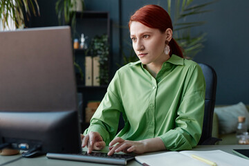 Portrait of smiling red haired young woman using computer at workplace in office decorated with...