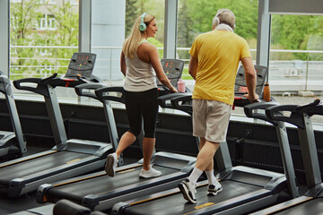 Fototapeta na wymiar Cardio training to fat burning. Senior man and woman in headphones walking on treadmills, focused on their fitness. Concept of sport, active seniors in modern life, healthy lifestyle. Ad