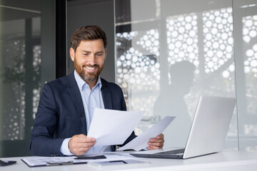 Portrait of a smiling young businessman sitting at an office desk with a laptop and looking through...