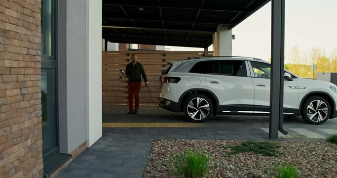 A man in casual clothes connects a charger to an electric car parked in front of a stylish cottage. A sunny day