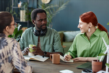 Portrait of adult African American man talking to colleagues sitting at meeting table in modern office with green plants