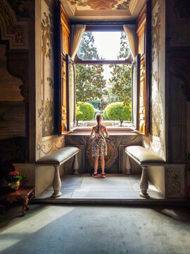 Rear view of a little girl looking through the open window of a room with a beautiful vintage interior.