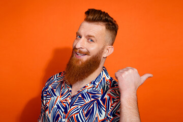 Photo of funky cheerful guy dressed print shirt pointing thumb back emtpy space isolated orange color background