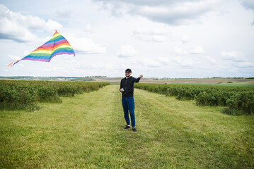 A young man wearing dark clothes holding a colorful rainbow kite in his hand. playing, standing in a lush green field. Summer spring leisure activities outdoors. Happiness in simple things