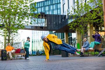 Young Stylish B-boy Breakdancing On Street Among Modern Buildings In Urban Area. Fashionable Group...