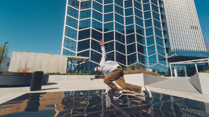 Outdoor Scene Capturing A Young Male Breakdancer Performing An Acrobatic Move In Urban Plaza, With Reflective Surfaces And A High-rise Building Background, Showcasing Youth, Energy, And Urban Culture.