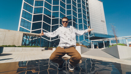 A Young Man Showcases His Breakdancing Skills In Front Of A Modern Glass Building, Under A Clear Blue Sky. His Dynamic Pose And Casual Attire Reflect A Vibrant Urban Youth Culture, Energetic Lifestyle