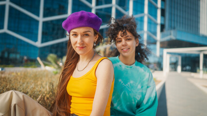 Vibrant Young Women In Colorful Attire Share A Joyful Moment In An Urban Setting, Their Smiles Radiating Friendship And Youthful Energy Against A Modern Architectural Background.