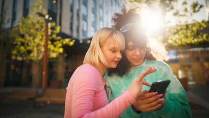 Two Young Women, One Blonde And One With Curly Hair, Joyfully Interact Over A Smartphone In A Sunlit Urban Setting, Embodying Friendship And The Digital Age Connection.