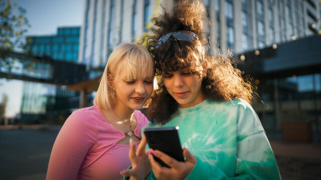 Two Young Women, One With Blonde Hair And The Other With Curly Brown Hair, Are Engrossed In A Smartphone Outdoors, Showcasing A Moment Of Connection And Digital Interaction In An Urban Setting.