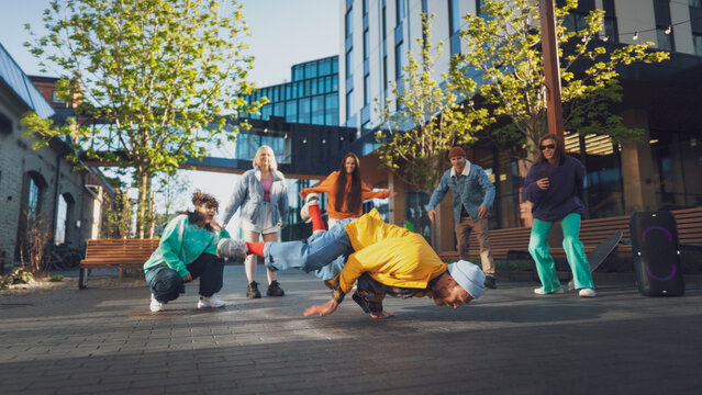 Vibrant Outdoor Scene Capturing A Diverse Group Of Young Adults Energetically Breakdancing In An Urban Setting, Surrounded By Modern Architecture And Greenery, Showcasing Youth, Culture, Friendship.