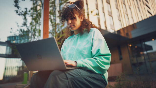 Young Woman With Curly Hair, Wearing A Tie-dye Sweatshirt, Works Intently On A Laptop Outdoors, Showcasing A Blend Of Technology, Education, And Casual Urban Lifestyle.