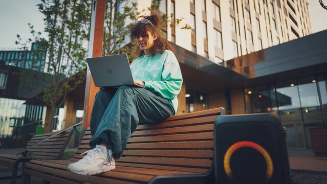 A Young Woman Enjoys Working On Her Laptop Outdoors, Seated On A Bench In An Urban Park At Dusk, Showcasing A Blend Of Technology, Casual Fashion, And Modern Lifestyle.