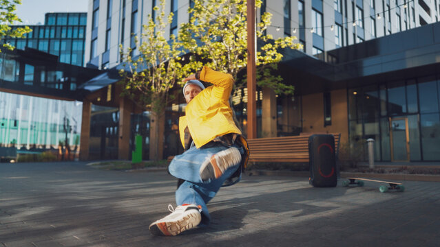 Dynamic Outdoor Scene Capturing A Young Male Breakdancer In Mid-air, Performing An Energetic Move In An Urban Setting With Modern Buildings And A Skateboard In The Background. Youth And Street Culture