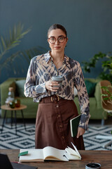 Vertical portrait of young confident businesswoman wearing tie dye blouse looking at camera standing in office interior with coffee cup