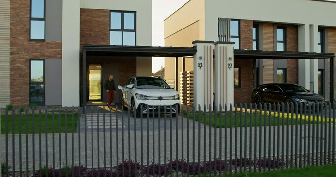 A man in casual clothes disconnects charging from an electric car parked in front of a stylish cottage. A sunny day