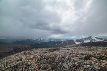 Dramatic top view from high stony pass to alpine valley against large mountain range with snow-capped peaks in gray rainy low clouds. Beautiful big snowy mountain tops in rain under grey cloudy sky. - 795433295