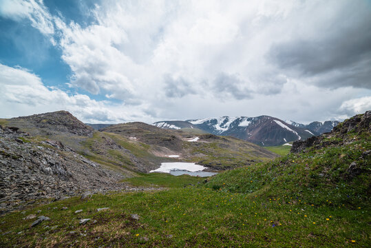 Beautiful small lake with snowfield among green grassy hills and rocks with view to snow-capped mountain range. Alpine lake among flowering meadows in high mountains. Blooming flowers in highlands.