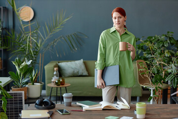 Portrait of young red haired businesswoman looking at camera standing in colorful office interior with coffee mug copy space