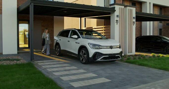 A woman in casual clothes disconnects charging from an electric car parked in front of a stylish cottage. A sunny day