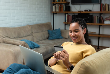 Young smiling African American woman holding credit card using laptop. Paying online, home shopping, e-commerce, internet banking, spending money, finance, electronic store concept from chair.