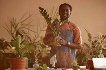 Waist up portrait of smiling African American man holding potted plant while enjoying gardening at home tinted peach