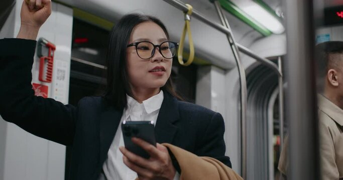 Frontal view of stylish Chinese girl holding on to safety handle inside moving bus or train. Checking on news or new posts in her smartphone. Beginning to jiggle with joy after reading something funny