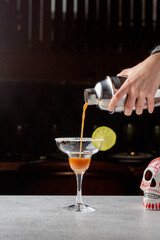 A professional bartender skillfully pours a vibrant red cocktail into a martini glass, garnished with a slice of lime, against a dark bar background