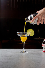 A professional bartender skillfully pours a vibrant red cocktail into a martini glass, garnished with a slice of lime, against a dark bar background