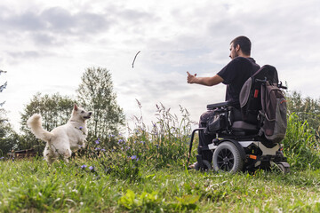 Man with physical impairment in a wheelchair and his best friend a beautiful white dog, enjoying a day outdoors. Pet love concept.