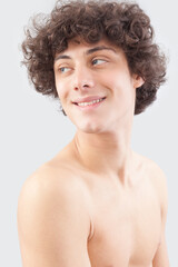 Smiling and handsome, a young man with curly hair, he looks to the side with his blue eyes, a portrait of a guy with a bare chest isolated against a gray background