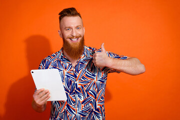 Portrait of toothy beaming man with red long beard hold tablet showing thumb up nice job isolated on vibrant orange color background