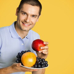 Square image - happy smiling man wear blue casual clothing, with plate of fruits, posing at studio against orange yellow color wall background.