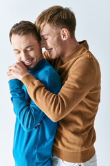 Obraz premium Two men in casual attire embrace each other in a loving hug against a gray backdrop.