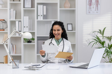 A cheerful Hispanic female doctor, in a white coat, reads an exciting letter or notification in a...